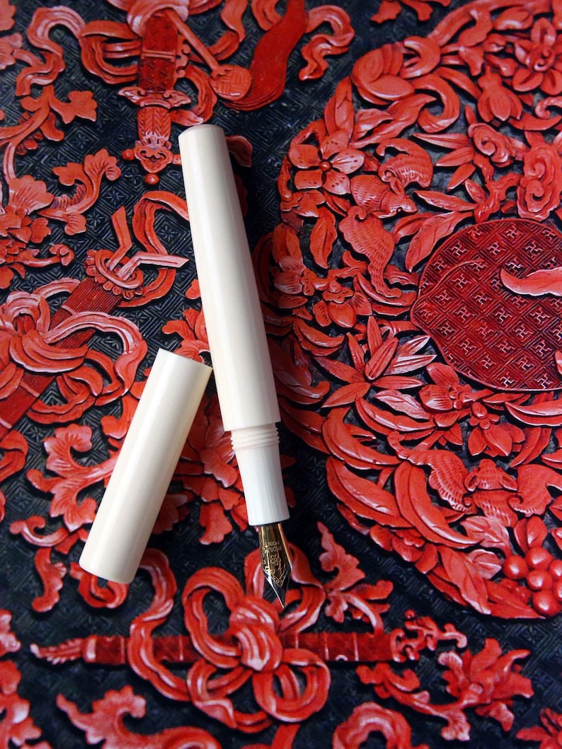 May include: A white fountain pen with a gold nib, resting on a red and black patterned surface. The pen is made of two sections that screw together.