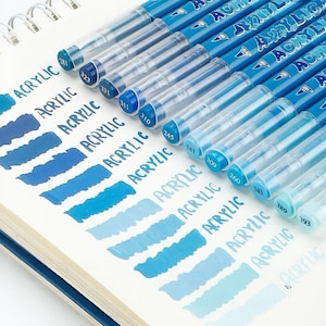 Puede incluir: Un conjunto de bolígrafos de pintura acrílica azules con tapas transparentes, dispuestos sobre un cuaderno blanco con espiral. Cada bolígrafo tiene un tono de azul diferente, con muestras de color correspondientes. La palabra "ACRYLIC" está impresa en los bolígrafos.