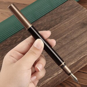 May include: A black and gold fountain pen with a gold nib is held in a hand. The pen has a shiny black barrel and a gold-colored cap and accents. The pen is on a wooden surface.