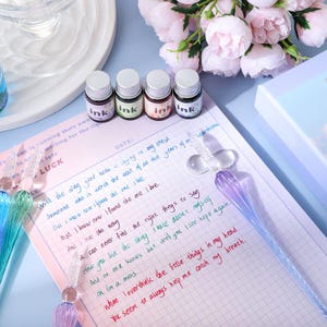 K&ouml;nnte beinhalten: Ein Schreibwarenset mit vier kleinen Tintenflaschen mit silbernen Deckeln, beschriftet mit "ink", in verschiedenen Farben. Ein Glas-Dip-Pen und ein Notizblock mit handschriftlichem Text sind ebenfalls vorhanden.