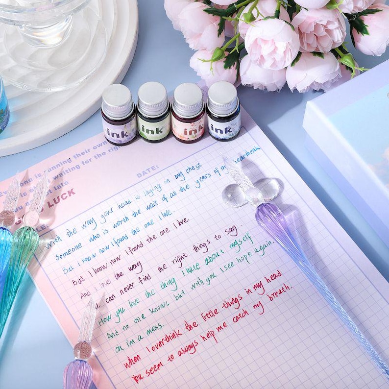 K&ouml;nnte beinhalten: Ein Schreibwarenset mit vier kleinen Tintenflaschen mit silbernen Deckeln, beschriftet mit "ink", in verschiedenen Farben. Ein Glas-Dip-Pen und ein Notizblock mit handschriftlichem Text sind ebenfalls vorhanden.