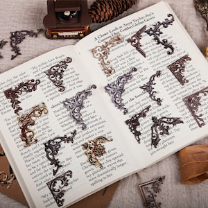 May include: Ornate metal corner embellishments in various metallic finishes, including bronze, silver, and gold, are arranged on an open book. The decorative elements feature intricate scrollwork and floral designs, ideal for scrapbooking or crafting.