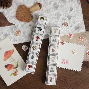 Op de afbeelding: Twee verticale stroken met witte vierkante rubberen stempels met diverse paddenstoelillustraties in bruine, rode en zwarte inkt. De stempels gaan vergezeld van diverse kleine paddenstoelstickers en een kaart met een gedetailleerde tekening.