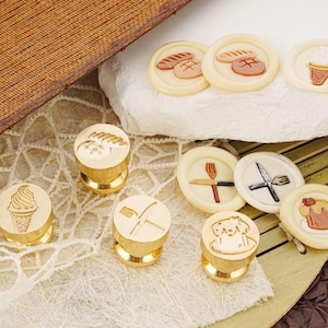 May include: A collection of gold-colored wax seal stamps with various food-related designs, including an ice cream cone, a fork and knife, and a cake. Several wax seals with bread and dessert designs are also visible.