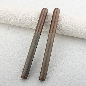 Puede incluir: Dos bol&iacute;grafos de metal color bronce con un cuerpo inferior acanalado y una parte superior lisa y redondeada. Los bol&iacute;grafos se muestran sobre una superficie blanca texturizada. Los bol&iacute;grafos tienen un aspecto vintage o antiguo.