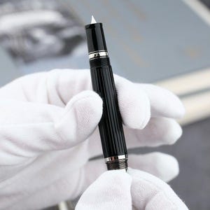 May include: A black fountain pen with silver detailing and a white nib. The pen features a ribbed barrel design. The pen is held by a person wearing white gloves. A writing instrument.