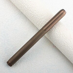 Puede incluir: Un bol&iacute;grafo de color bronce con un dise&ntilde;o acanalado a lo largo del cuerpo. El bol&iacute;grafo tiene forma cil&iacute;ndrica con una punta ligeramente redondeada y un extremo roscado. El bol&iacute;grafo est&aacute; sobre un fondo blanco texturizado.