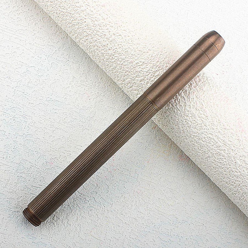 Puede incluir: Un bol&iacute;grafo de color bronce con un dise&ntilde;o acanalado a lo largo del cuerpo. El bol&iacute;grafo tiene forma cil&iacute;ndrica con una punta ligeramente redondeada y un extremo roscado. El bol&iacute;grafo est&aacute; sobre un fondo blanco texturizado.