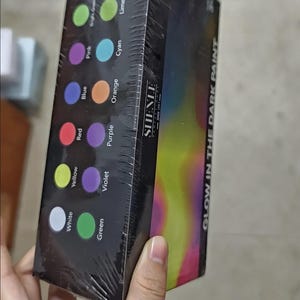 Puede incluir: Una caja rectangular de pintura que brilla en la oscuridad con 12 colores. El empaque negro muestra la marca SHENLE, el texto GLOW IN THE DARK PAINT y muestras circulares etiquetadas como White, Yellow, Red, Blue, Pink, Bright Green, Green, Violet, Purple, Orange, Cyan y Lime.