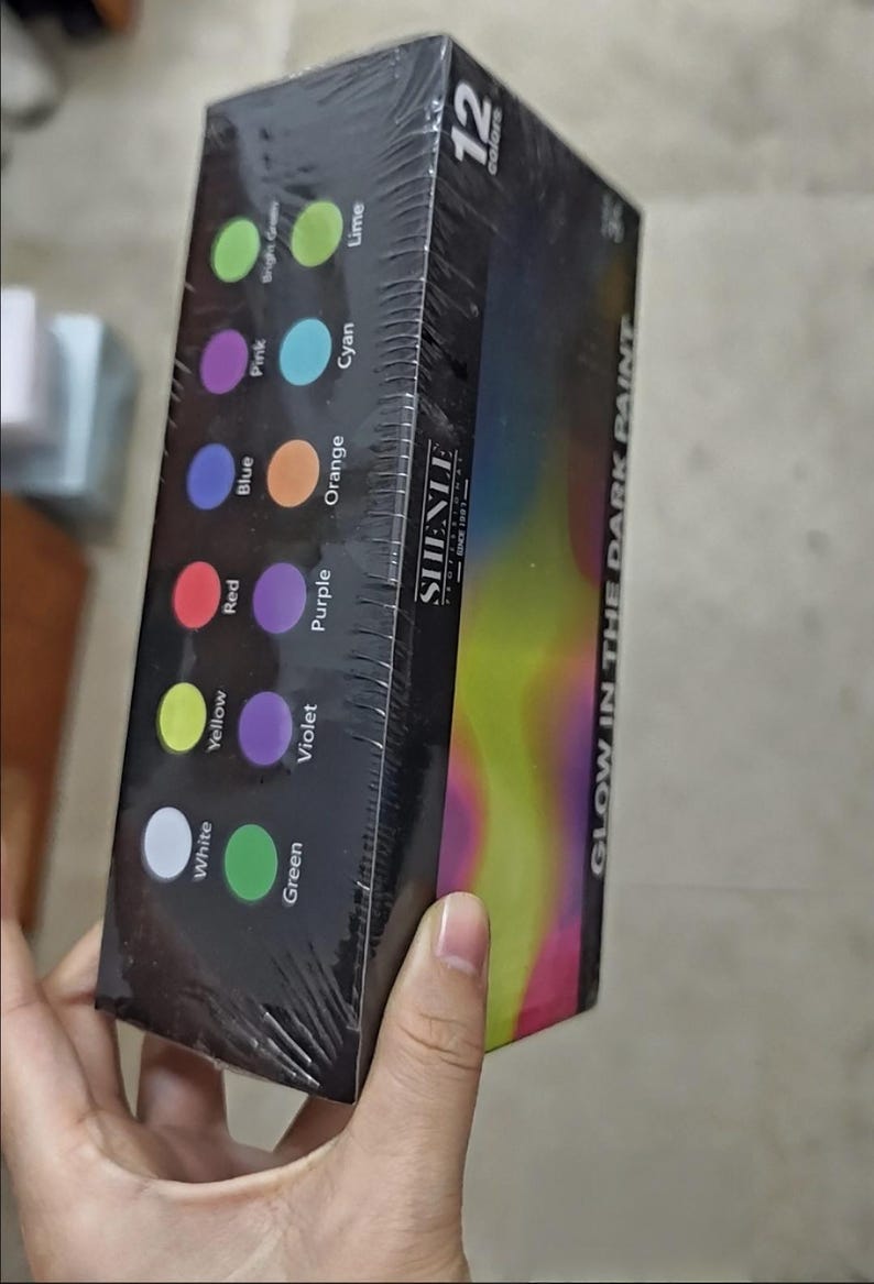Puede incluir: Una caja rectangular de pintura que brilla en la oscuridad con 12 colores. El empaque negro muestra la marca SHENLE, el texto GLOW IN THE DARK PAINT y muestras circulares etiquetadas como White, Yellow, Red, Blue, Pink, Bright Green, Green, Violet, Purple, Orange, Cyan y Lime.