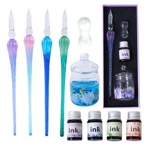 K&ouml;nnte beinhalten: Ein Kalligraphie-Set mit Glas-Dip-Pens in Lila, Rosa, Blau und Gr&uuml;n. Das Set enth&auml;lt Tintenflaschen, ein Glas-Tintenfass und ein dekoratives Glas mit blauer Fl&uuml;ssigkeit und floralen Akzenten. Die Tintenflaschen sind mit dem Wort "ink" beschriftet.