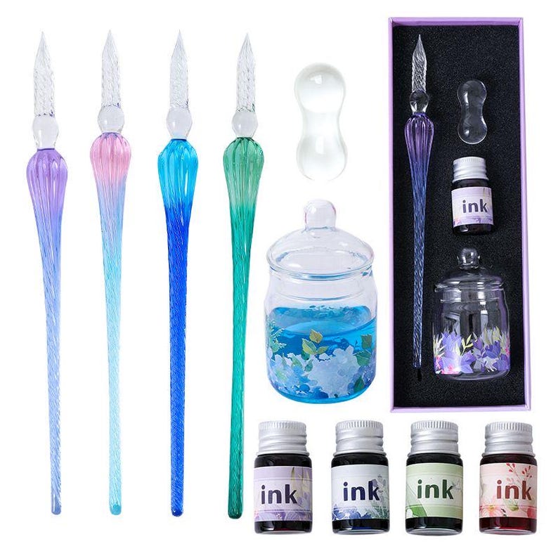 K&ouml;nnte beinhalten: Ein Kalligraphie-Set mit Glas-Dip-Pens in Lila, Rosa, Blau und Gr&uuml;n. Das Set enth&auml;lt Tintenflaschen, ein Glas-Tintenfass und ein dekoratives Glas mit blauer Fl&uuml;ssigkeit und floralen Akzenten. Die Tintenflaschen sind mit dem Wort "ink" beschriftet.