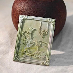 May include: A rectangular, light green relief carving with a classical still life scene. The carving features a decorative pitcher, a wine glass, and a plate with fruit, all within a detailed frame. The background is a dark brown, possibly wooden, sphere.