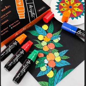 May include: A set of black acrylic markers with colored caps, including orange, red, green, and blue. The packaging box displays the text Acrylic marker, Water Based, and Safe To Use. A black sheet features a hand-drawn illustration of fruit.
