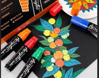 May include: A set of black acrylic markers with colored caps, including orange, red, green, and blue. The packaging box displays the text Acrylic marker, Water Based, and Safe To Use. A black sheet features a hand-drawn illustration of fruit.
