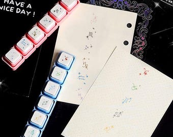 May include: Two plastic keycap sets in pink and blue, each featuring six white keys with colorful zodiac symbols. Two cream-colored paper sheets are included, one with two binder holes, both printed with vertical rows of zodiac signs and small stars.