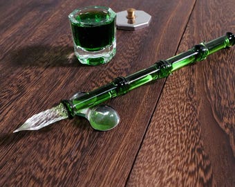 May include: A green glass dip pen with a bamboo-like texture and a clear, spiraled glass nib, accompanied by a small, square glass inkwell filled with dark green ink and a separate metallic, octagonal lid with a small central knob.
