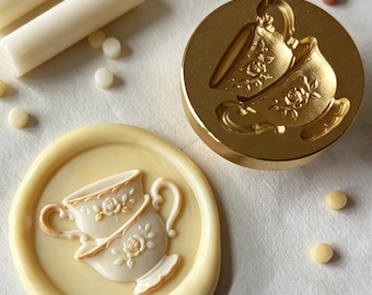 May include: A circular gold-toned metal wax seal stamp featuring an engraved design of two stacked teacups with floral details, alongside a cream-colored wax seal impression of the same design and several small wax sticks in cream and tan colors.