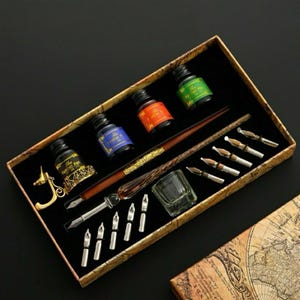 Op de afbeelding: Een kalligrafieset in een doos met kaartpatroon, bestaande uit een bruine houten pen met een goudkleurige metalen greep, drie glazen inktpotjes met het label The Feather Pen in zwart, blauw en groen, een goudkleurige pennenhouder, een klein glazen bakje en tien metalen penpunten.