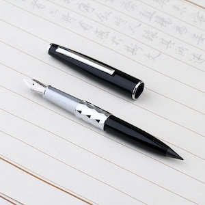 May include: A black and silver fountain pen, cap removed, on lined paper. The pen has a silver nib and a geometric barrel design. The cap is black with a silver clip. The paper has writing.