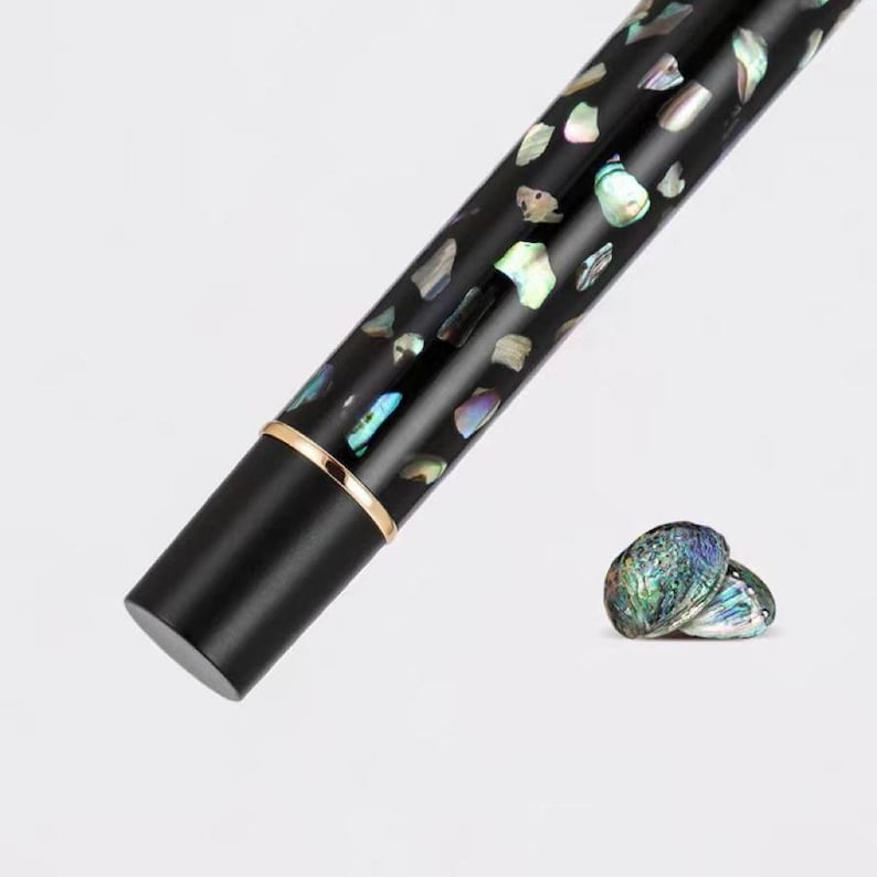 Puede incluir: Un bol&iacute;grafo negro con incrustaciones iridiscentes en forma de concha y una banda dorada. Dos conchas de abul&oacute;n est&aacute;n junto al bol&iacute;grafo. El dise&ntilde;o del bol&iacute;grafo presenta un acabado brillante y las incrustaciones de concha tienen una variedad de colores.