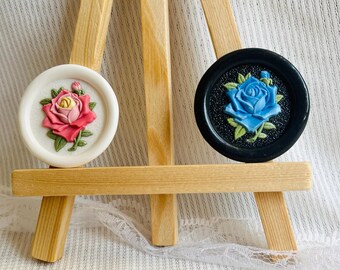 May include: Two circular decorative buttons featuring floral designs. One button is white with a pink and yellow rose. The other button is black with a blue rose. Both buttons have detailed green leaves and are displayed on a wooden easel.