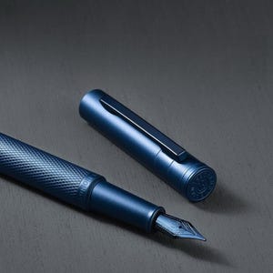 Op de afbeelding: Een blauwe vulpen met een getextureerde behuizing en een bijpassende dop. De dop is losgemaakt, waardoor de penpunt zichtbaar is. De pen heeft een strak, modern ontwerp tegen een donkere achtergrond.
