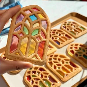 May include: A close-up of a stained-glass window cookie held in a hand, with several similar cookies on a baking sheet. The cookies are rectangular with a stained-glass design filled with colorful, translucent candy. The text "Monster & Sugar" is visible.