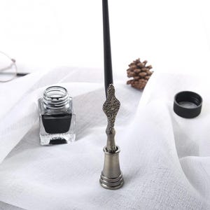 Op de afbeelding: Een vintage dip pen met een decoratieve metalen handgreep en een zwarte penpunt, rustend in een bijpassende inktpot. Een vierkante glazen inktfles gevuld met zwarte inkt staat in de buurt. Een zwarte dop en een dennenappel zijn ook zichtbaar.