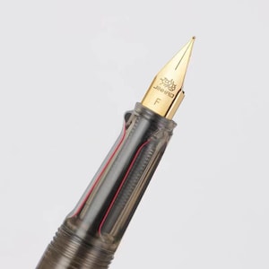 Puede incluir: Primer plano de una pluma estilogr&aacute;fica Jinhao. La pluma tiene un plum&iacute;n dorado con el nombre de la marca y la letra "F" grabados. El cuerpo de la pluma es gris transparente, revelando un componente interno rojo.