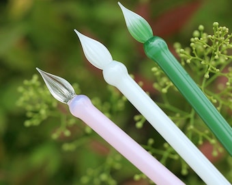 May include: Three glass dip pens with different colored glass bodies: pink, white, and green. The tips of the pens are all pointed and have a slight curve.