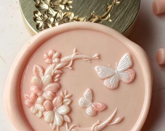 May include: A round, pale pink wax seal featuring a raised design of a flowering branch and two butterflies, positioned in front of a circular gold-toned metal stamp head with an identical engraved butterfly and floral pattern.