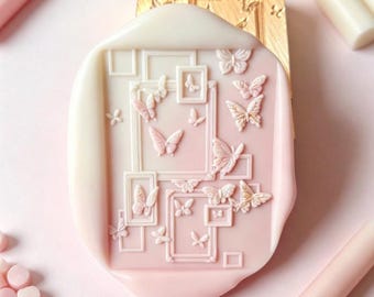 May include: A wax seal featuring a gradient of cream to light pink, embossed with a collection of rectangular frames and various butterfly shapes. The seal is positioned in front of a gold metal stamp head, with pink and white wax sticks nearby.