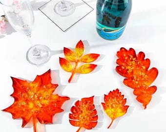 May include: A collection of five decorative resin leaf-shaped coasters and ornaments in shades of orange, red, and yellow, featuring embedded gold flakes. The set includes various maple and oak leaf designs alongside a clear glass and a bottle.
