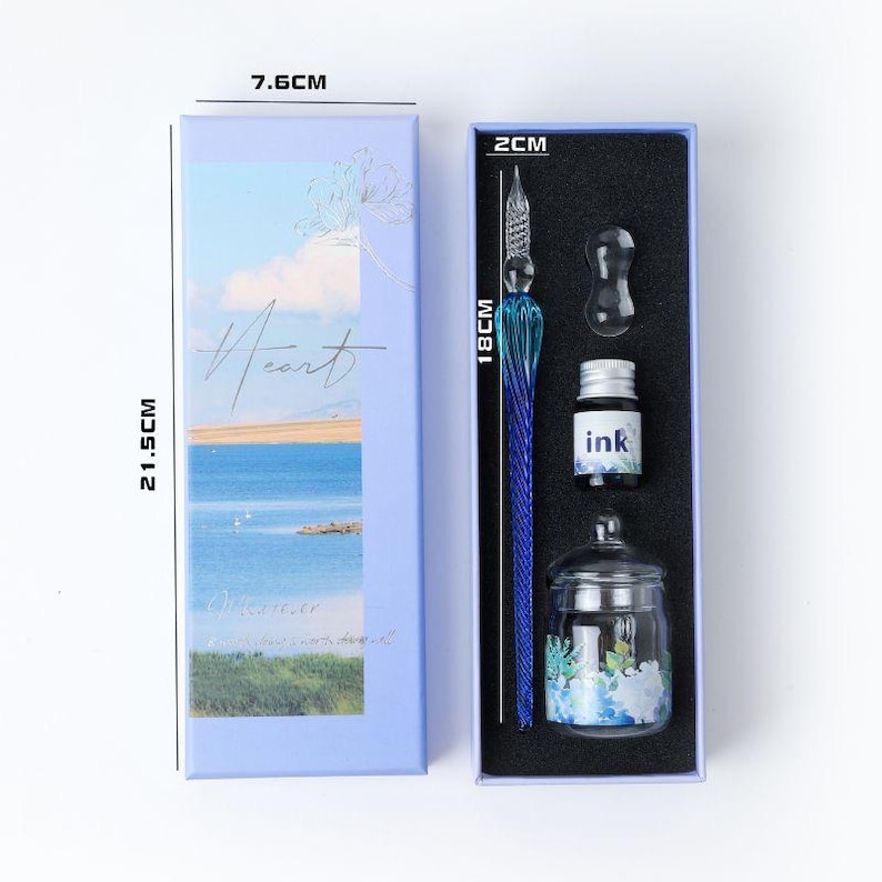 K&ouml;nnte beinhalten: Ein Kalligraphie-Set in einer hellblauen Box mit einem blauen Glasstift (18 cm), Tinte und einem Glas-Tintenfass. Die Box hat ein Landschaftsdesign mit dem Wort "Heart" auf der Vorderseite. Die Box misst 21,5 cm x 7,6 cm.