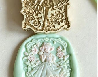 May include: A rectangular gold-toned metal wax seal stamp featuring an embossed figure in a dress surrounded by flowers, paired with a light green wax impression showing the same detailed design of a person standing amidst floral patterns.
