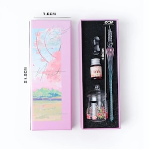 K&ouml;nnte beinhalten: Rosa Geschenkbox mit Glasfeder, Tintenfass, Tintenf&auml;sschen und Federhalter. Der Stift ist 18 cm lang. Die Box ist 21,5 cm hoch und 7,6 cm breit. Die Box hat eine Aquarellillustration eines Riesenrads und Kirschbl&uuml;ten.