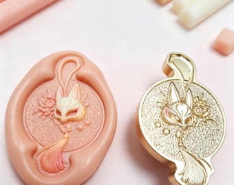 May include: A peach-colored silicone mold and a matching gold-toned metal wax seal stamp. Both items feature an embossed design of a fox mask with a tassel and floral accents. Several wax sticks in peach, gold, and white are arranged nearby.