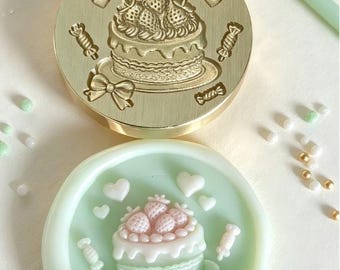 May include: A circular brass wax seal stamp featuring an engraved cake with strawberries, hearts, bows, and candies. Below it is a light green wax seal impression showing the same cake design in raised relief with pink and white details.