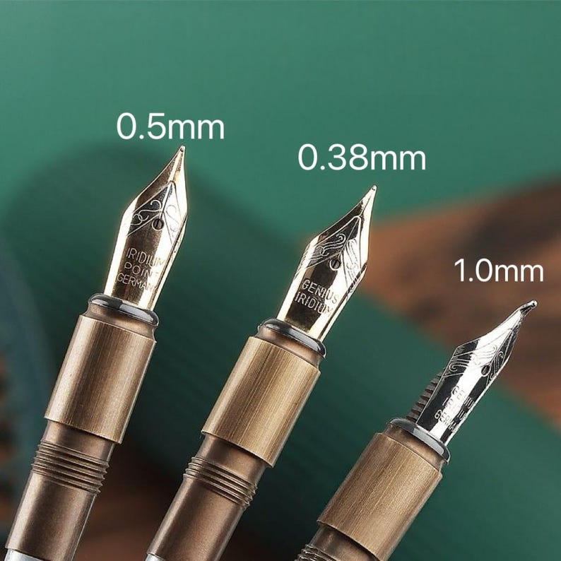 May include: Close-up of three fountain pen nibs with varying widths: 0.5mm, 0.38mm, and 1.0mm. The nibs are gold-colored with intricate designs and text. The pen bodies are a bronze color. The background is a dark green.