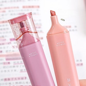May include: Two pastel-colored highlighters with the text "NEBULA COLOR" printed on the side. One is pink with a clear, glitter-filled cap, and the other is peach. The peach highlighter has a paperclip resting on top.
