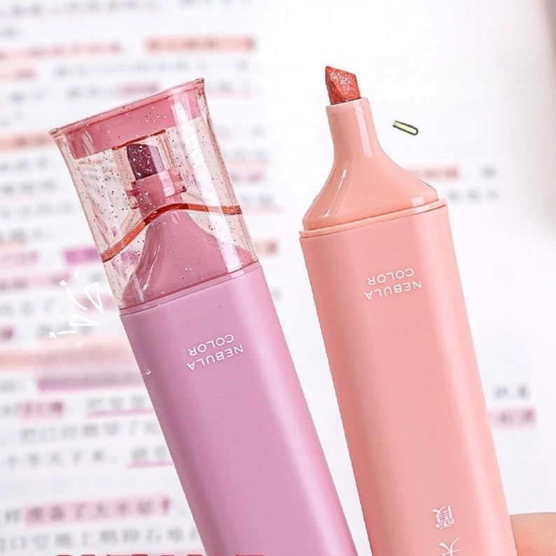 May include: Two pastel-colored highlighters with the text "NEBULA COLOR" printed on the side. One is pink with a clear, glitter-filled cap, and the other is peach. The peach highlighter has a paperclip resting on top.