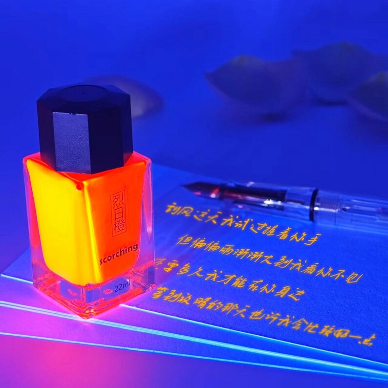May include: A clear glass bottle of "scorching" nail polish with a black cap, illuminated with yellow and orange hues. The bottle is next to a pen and a sheet of paper with Chinese characters.