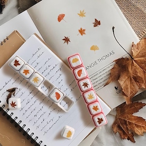 May include: Two sets of leaf-themed stamps in a fall setting. One set has a pink border, the other is clear. The stamps feature various leaf designs in shades of orange, yellow, and brown. The background includes a notebook, loose leaves, and text.