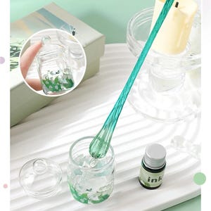 K&ouml;nnte beinhalten: Ein klares Glas mit Deckel, gef&uuml;llt mit einem floralen Design, und ein langer, gr&uuml;ner Glasr&uuml;hrstab. Eine kleine Tintenflasche mit wei&szlig;em Etikett ist ebenfalls vorhanden. Das Glas hat einen Durchmesser von etwa 5 cm.