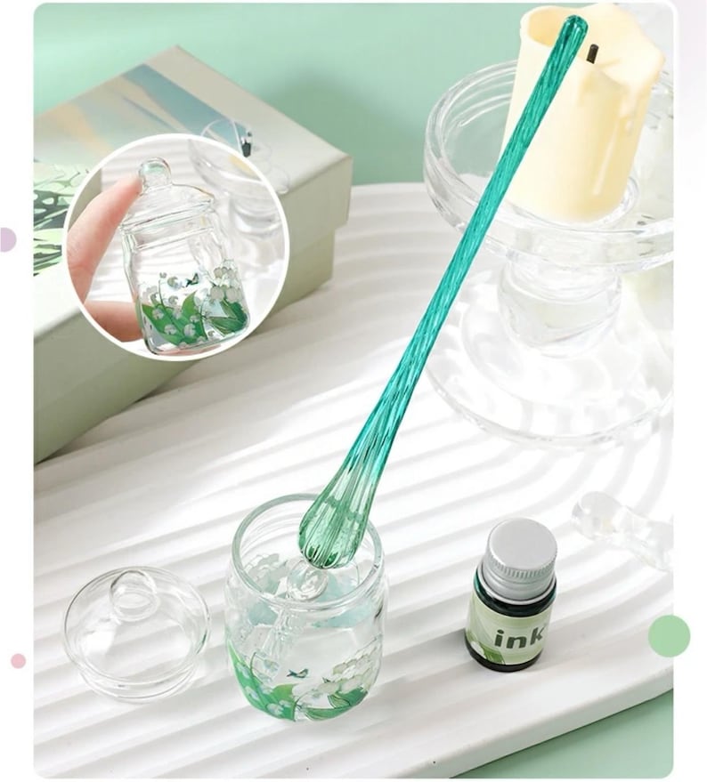 K&ouml;nnte beinhalten: Ein klares Glas mit Deckel, gef&uuml;llt mit einem floralen Design, und ein langer, gr&uuml;ner Glasr&uuml;hrstab. Eine kleine Tintenflasche mit wei&szlig;em Etikett ist ebenfalls vorhanden. Das Glas hat einen Durchmesser von etwa 5 cm.