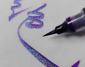 May include: A brush pen with a dark barrel and a fine tip is shown applying a vibrant, shimmering purple and blue glitter ink onto a white surface. The ink creates fluid, swirling lines that display a distinct color-shifting effect.