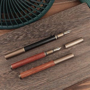 May include: Three fountain pens with metal accents and wooden barrels. One pen is black, and two are reddish-brown. The pens are displayed on a wooden surface, with one pen's cap removed, revealing the nib.