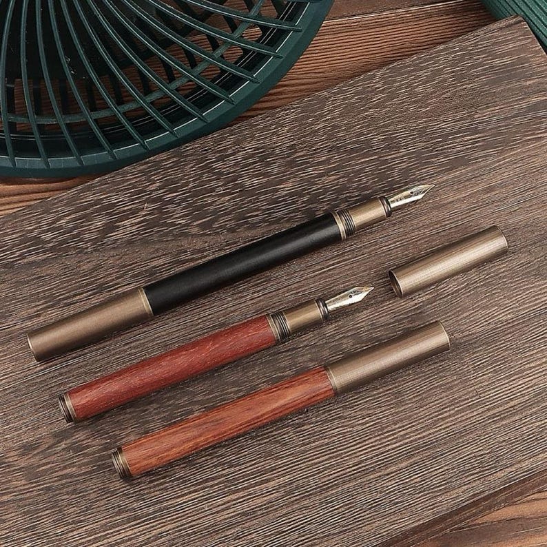 May include: Three fountain pens with metal accents and wooden barrels. One pen is black, and two are reddish-brown. The pens are displayed on a wooden surface, with one pen's cap removed, revealing the nib.