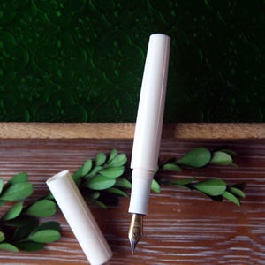 May include: A white fountain pen with a gold nib and a matching white cap. The pen is lying on a wooden surface with green foliage.